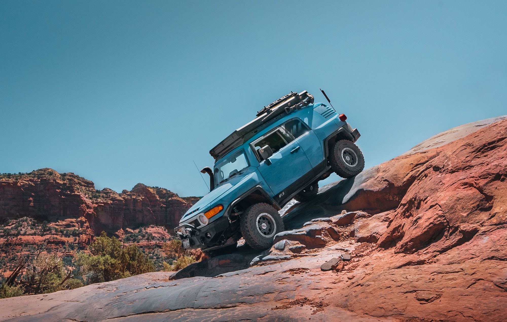 Blue Toyota FJ Cruiser climbing red slickrock, one rear wheel in the air